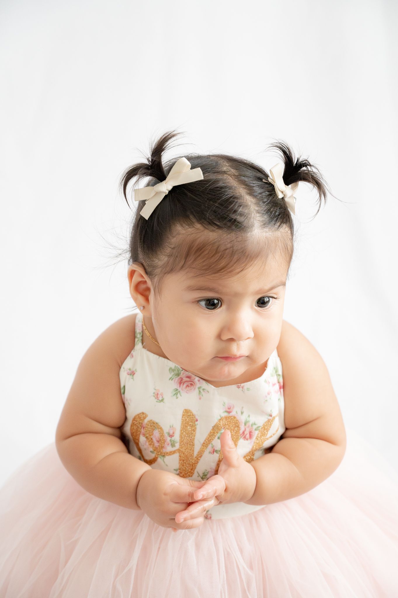 Baby wearing pink tulle dress and hair bows sits for portrait against white background.