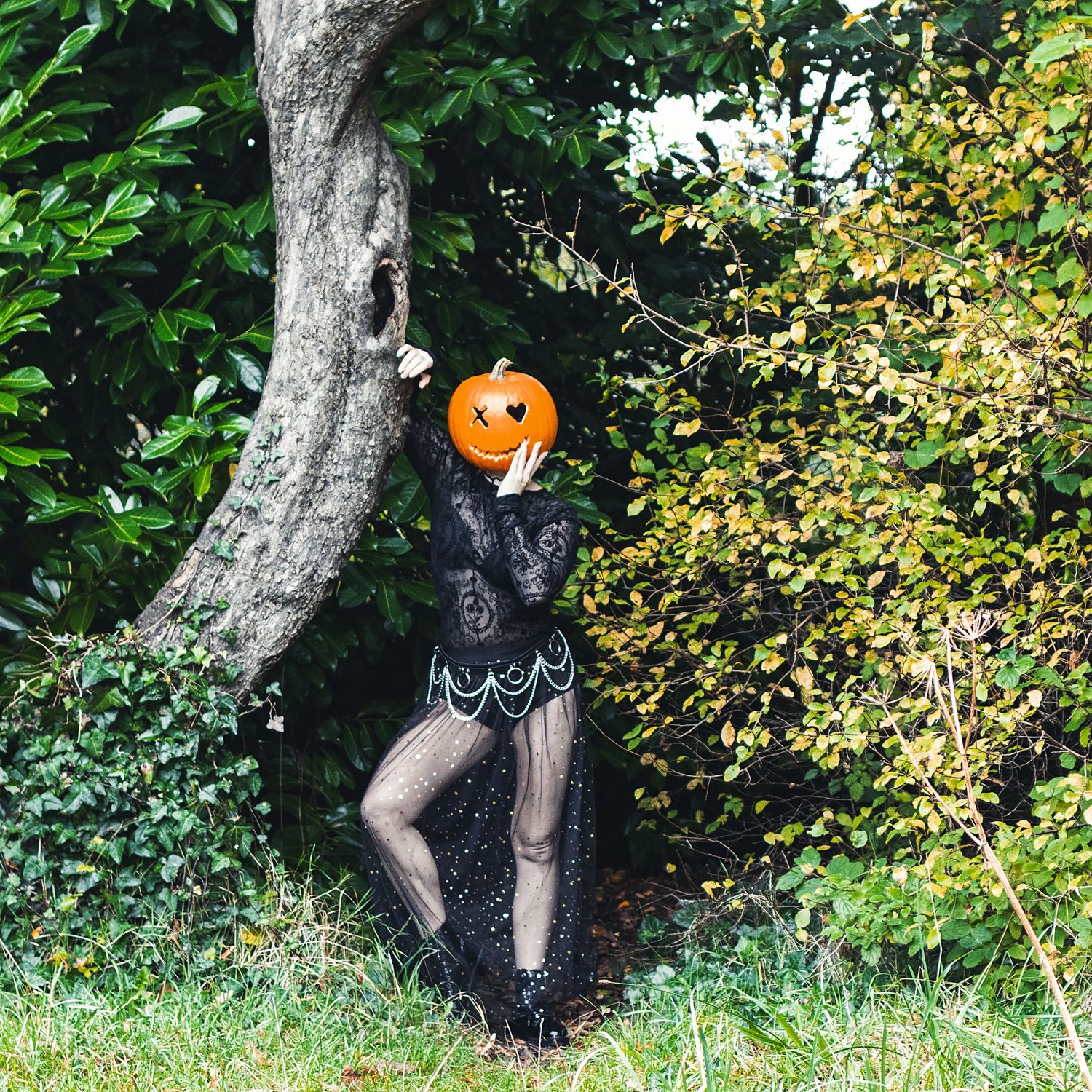 Spooky figure wearing black dress and pumpkin head poses against autumn foliage.