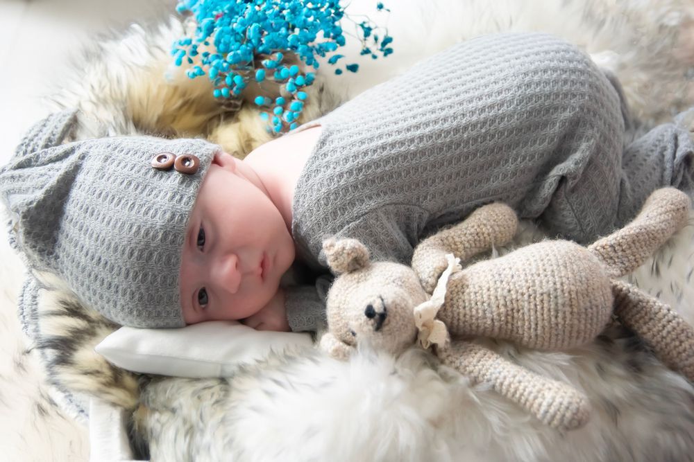 A small baby wearing a gray knit hat lies on a fur blanket next to plush toys and blue flowers.