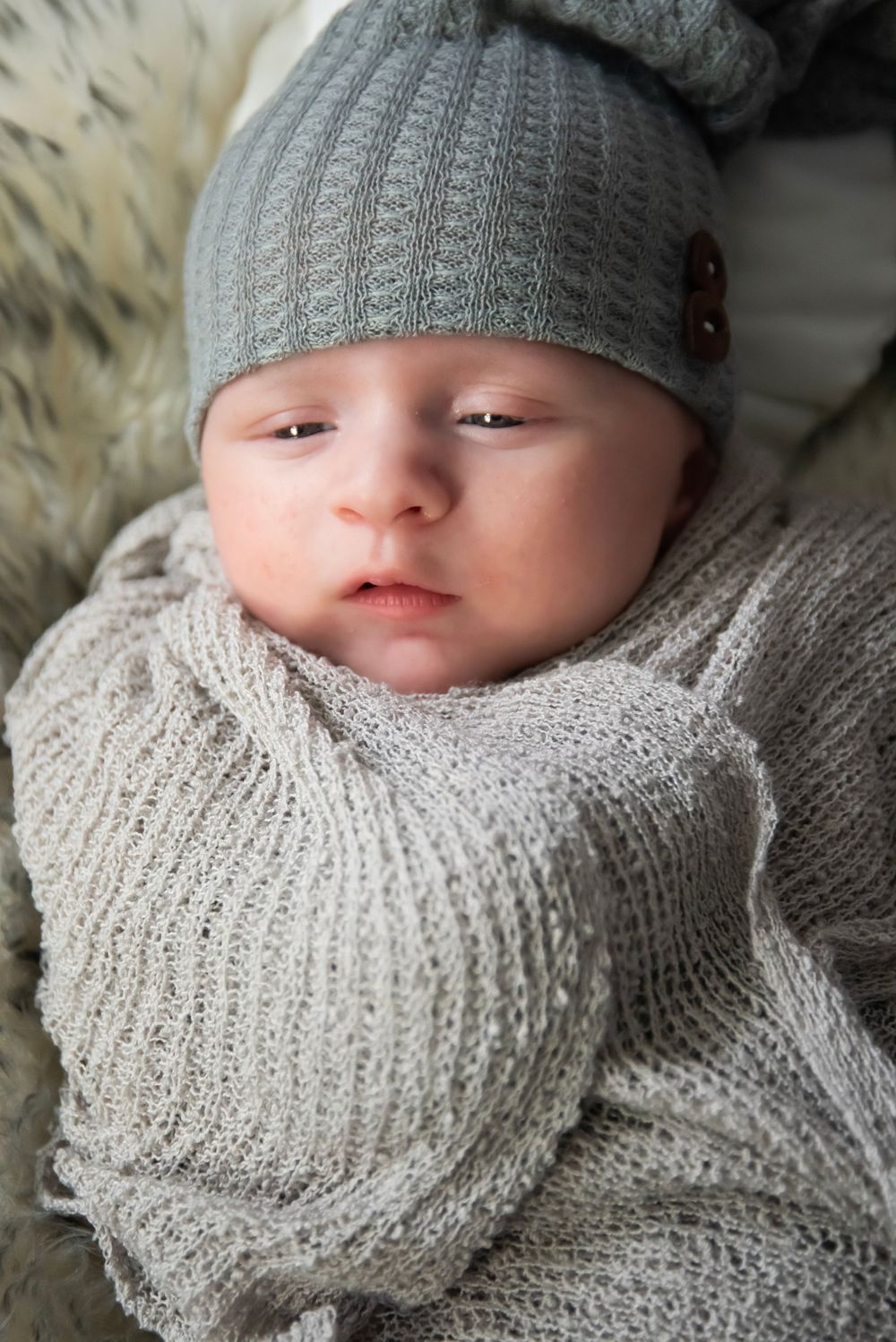 A bundled baby wearing a gray knitted beanie and cozy sweater rests peacefully on a soft furry surface.