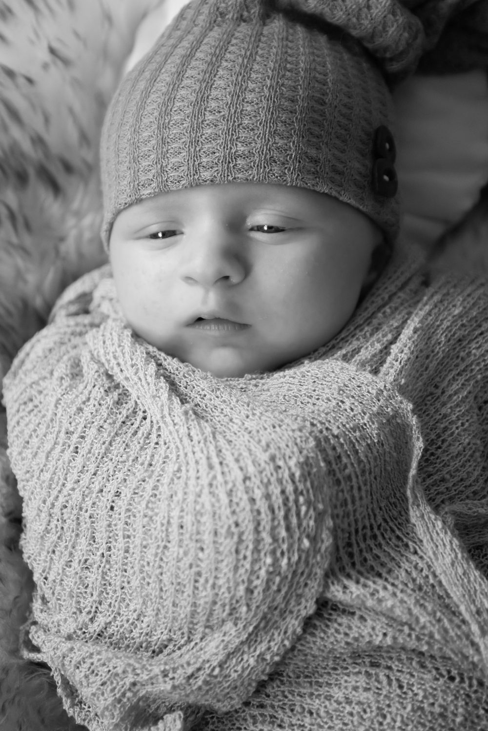 Black and white portrait of a tiny baby wearing a knitted cap and sweater looking cozy.
