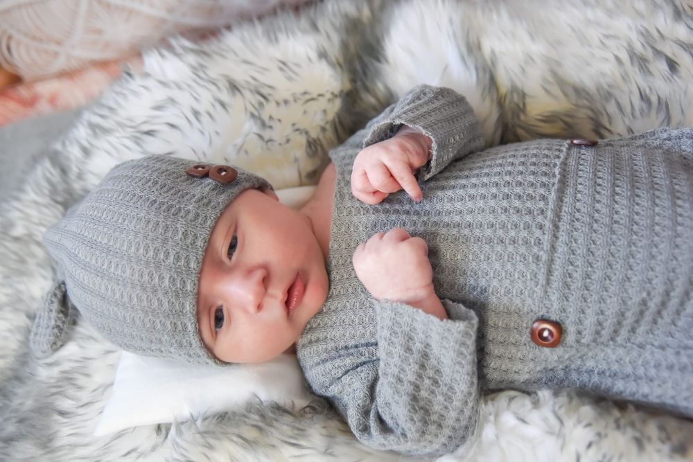 Newborn wearing a cozy gray knit sweater and bonnet lying on white bedding.