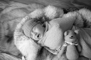 Black and white photograph of a sleeping baby in knitted sweater and soft blanket with a small plush toy.