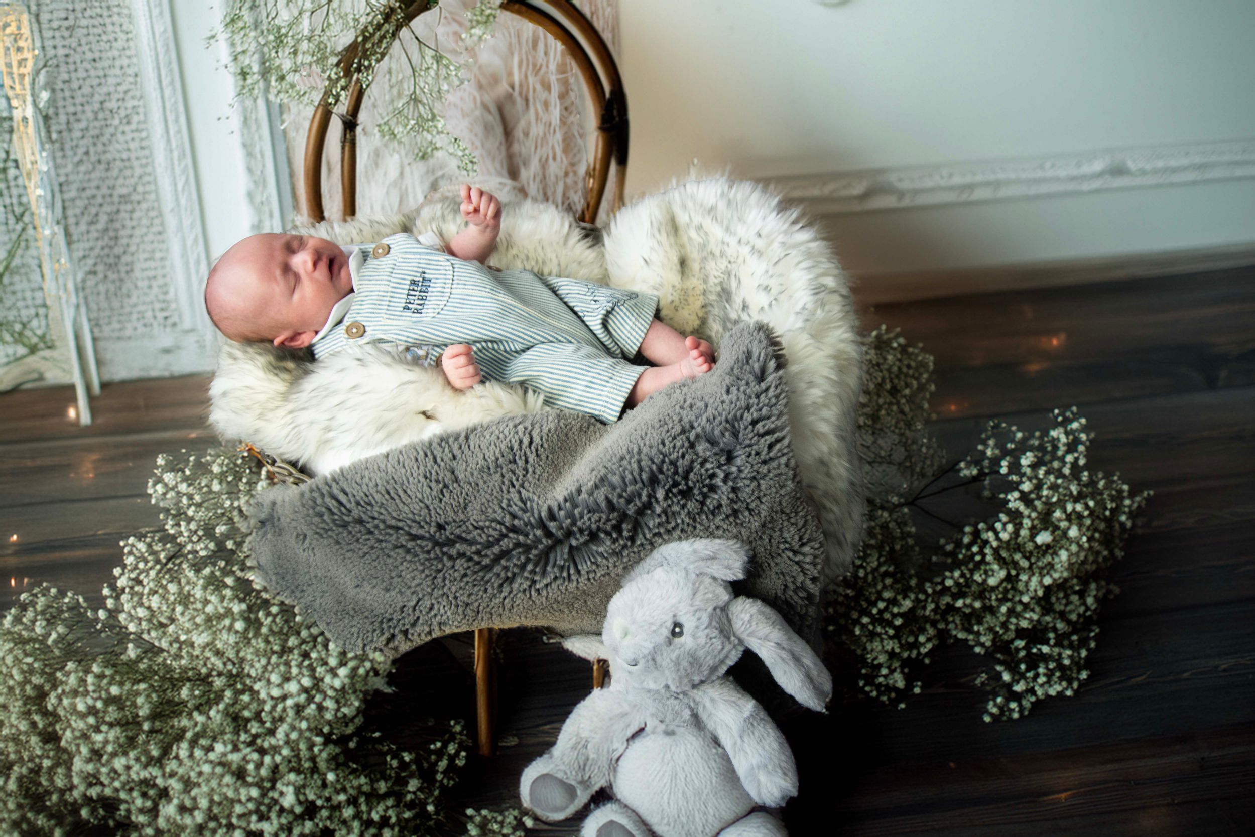 A baby lies on a gray blanket surrounded by greenery and a plush bunny toy on a wooden floor.