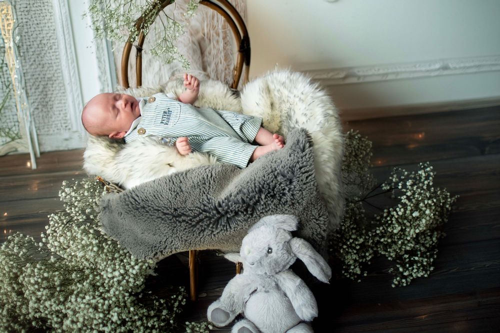 A baby lies on a gray blanket surrounded by greenery and a plush bunny toy on a wooden floor.