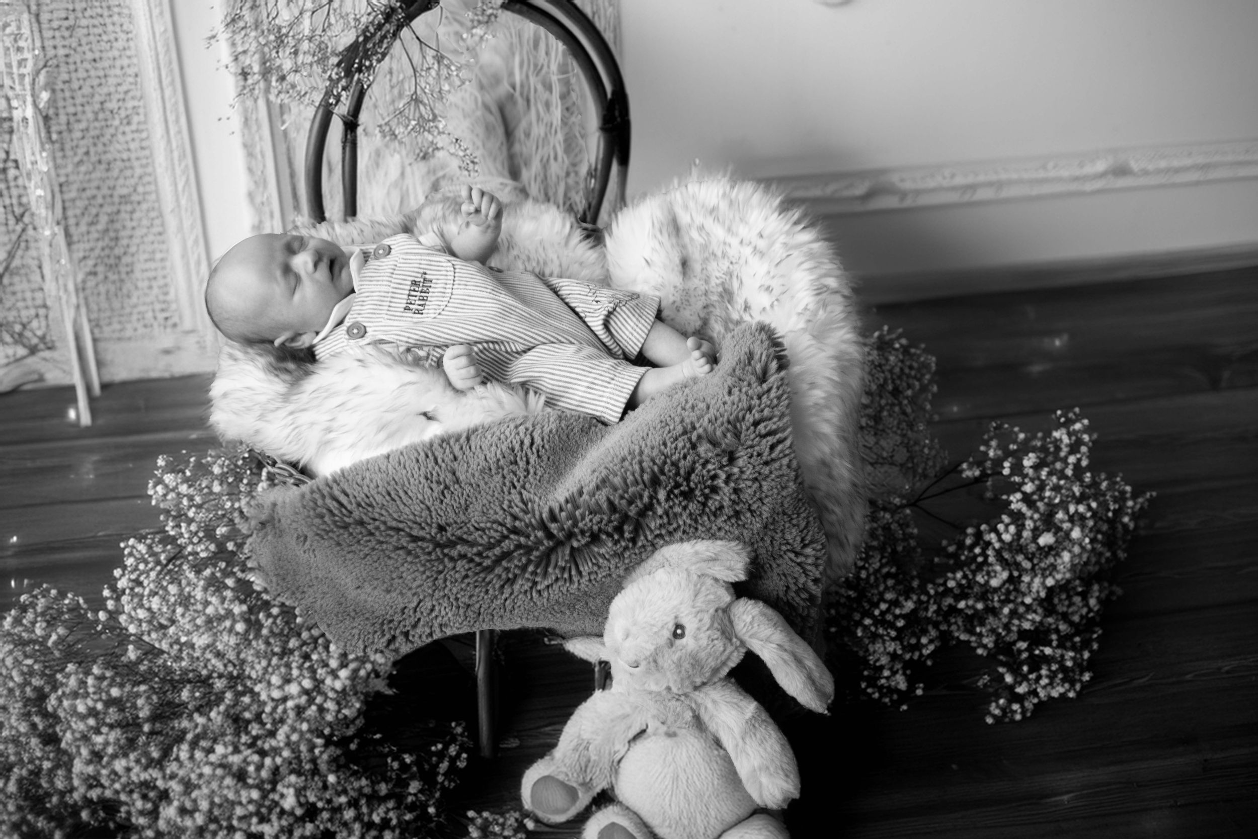 A vintage black and white photograph shows a baby sleeping in a bassinet surrounded by stuffed animals and flowers.