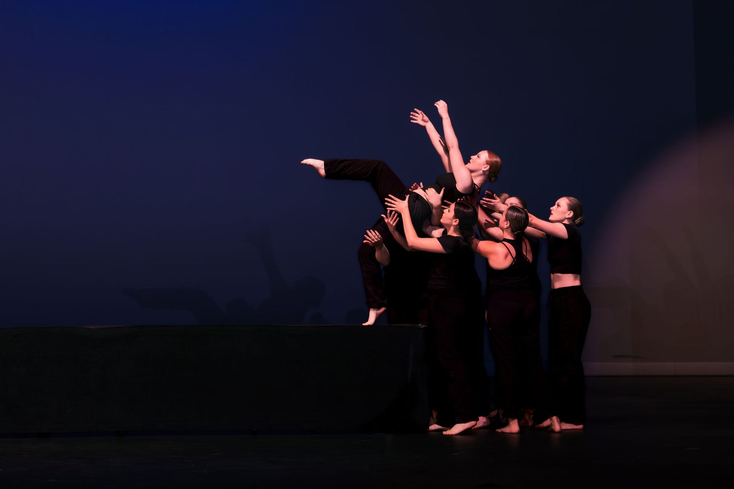 Dancers in black performing a contemporary dance piece on a dark stage with dramatic lighting and synchronized movements.
