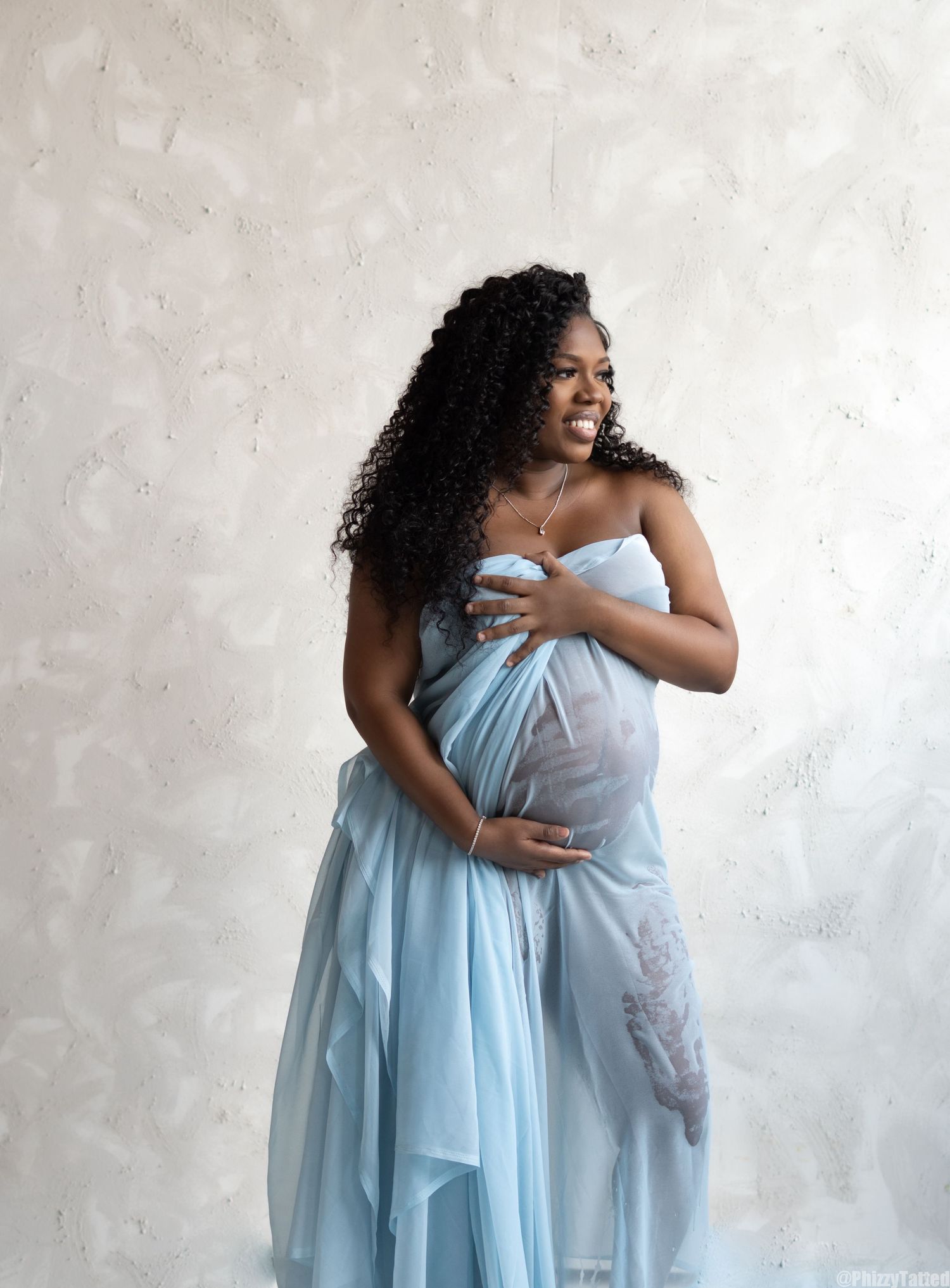 Maternity photoshoot featuring woman in flowing light blue gown against textured wall.