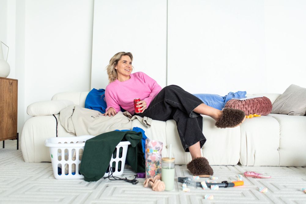 Person in pink sweater relaxes on white couch surrounded by laundry baskets and scattered clothes on floor.