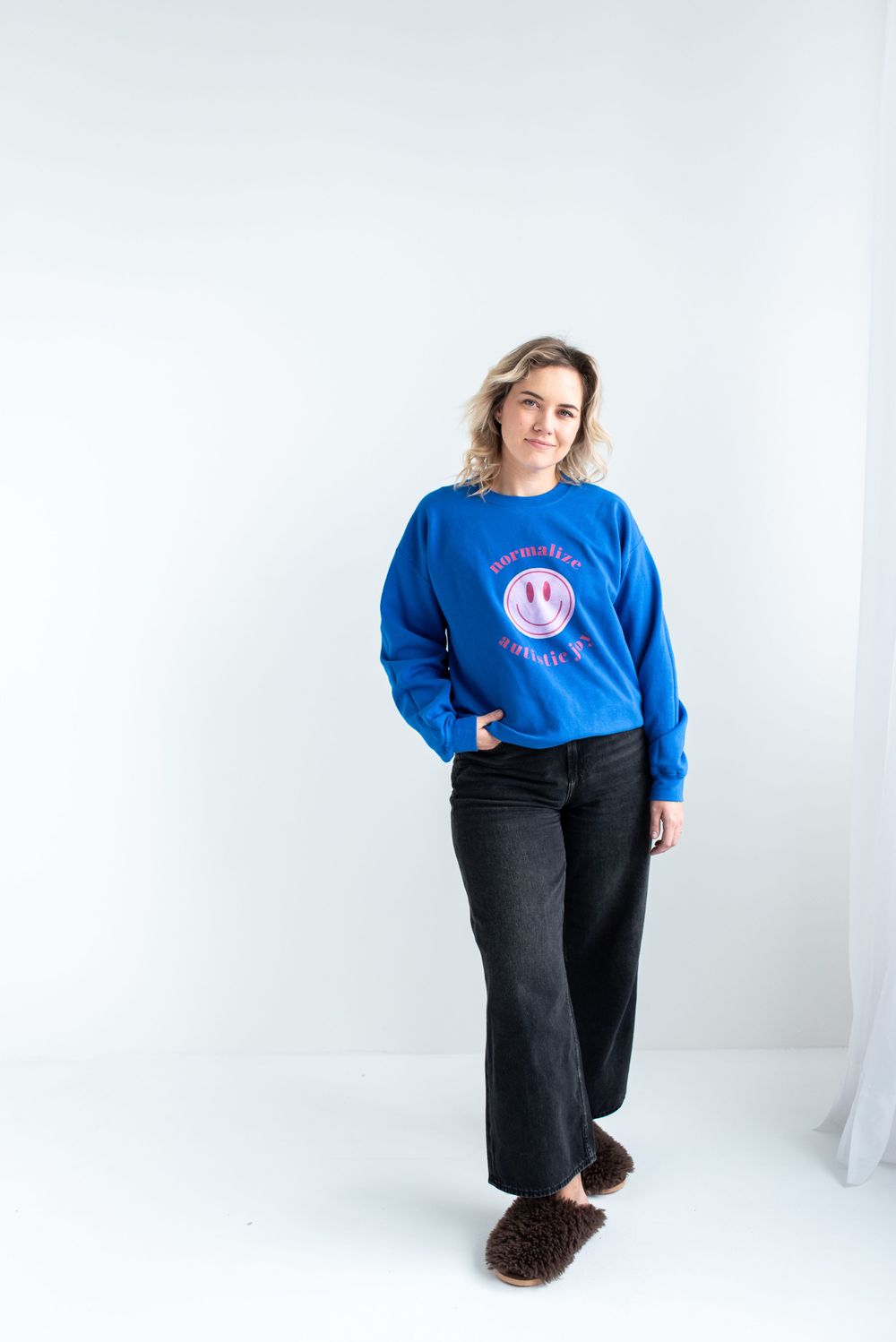 Someone wearing a bright blue sweatshirt with a smiley face design stands against a white background in dark jeans and slippers.