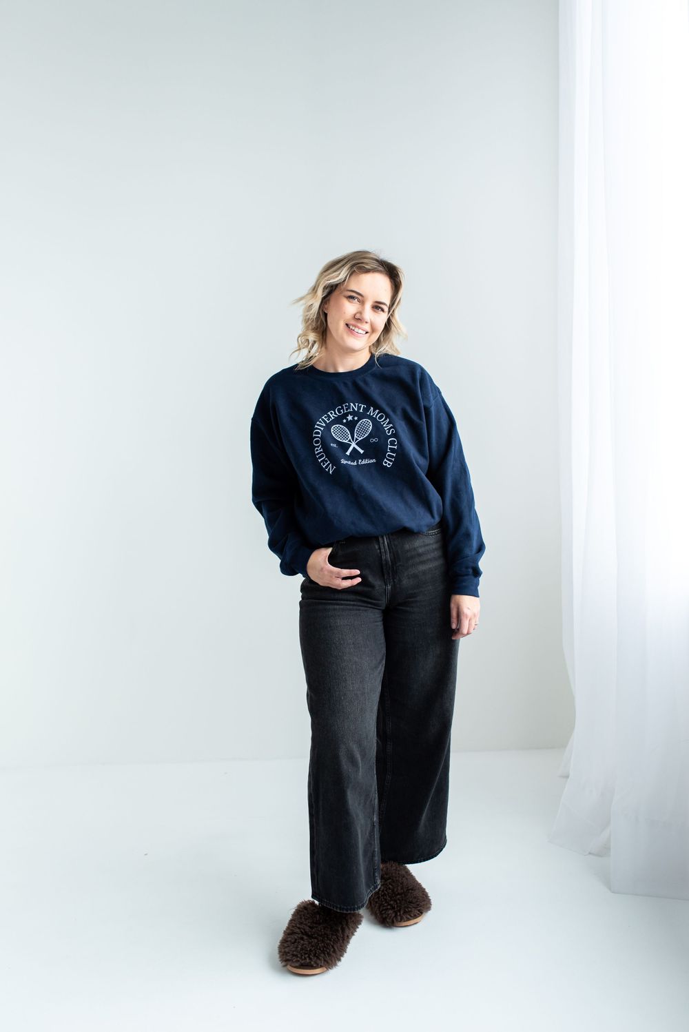 Person wearing a navy blue crewneck sweater with graphic logo stands against white background in dark jeans and brown shoes.
