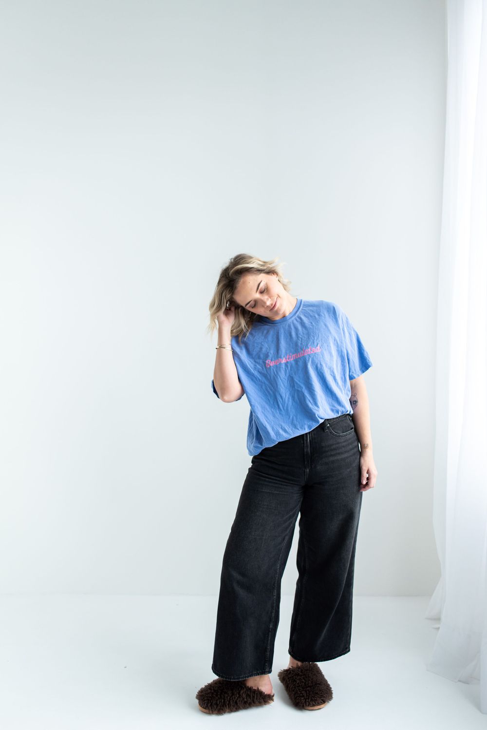A casual pose in a blue t-shirt and wide-leg black pants with fuzzy brown slippers against a white background.