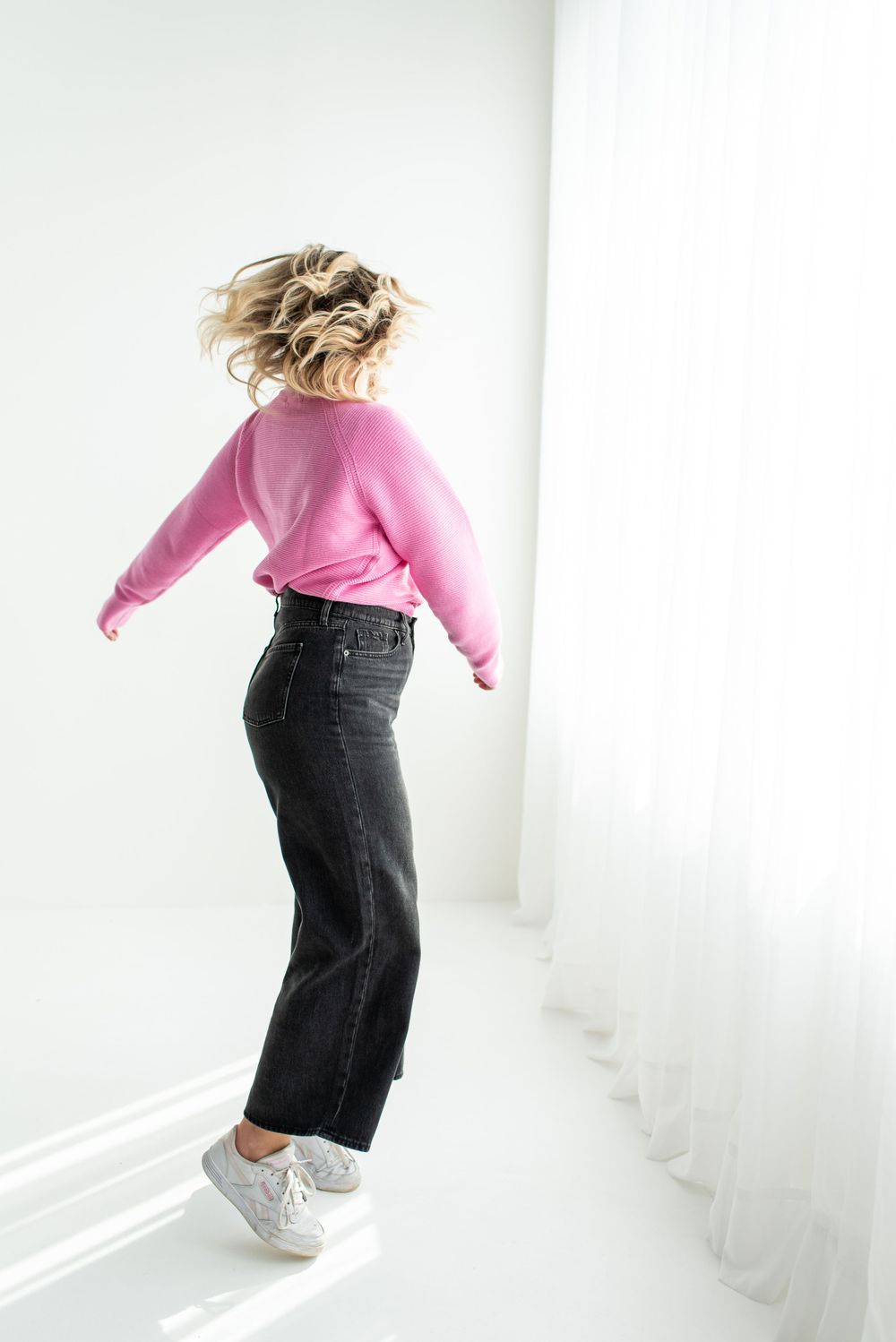 A person in a pink sweater and black jeans jumps against a white wall while wearing gray sneakers.