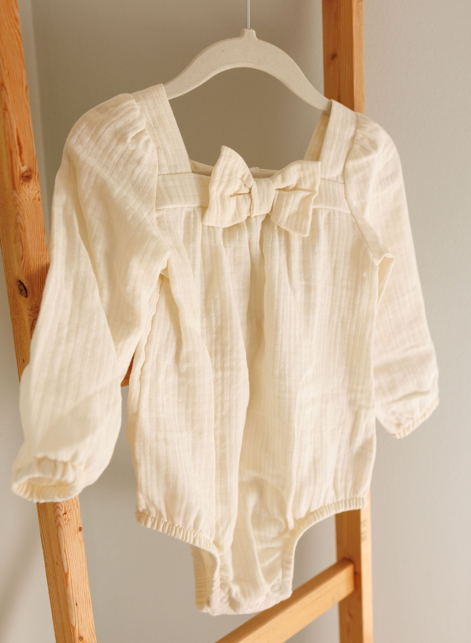 Cream-colored cotton bodysuit with long sleeves and bow detail hanging on white hanger against wooden ladder.