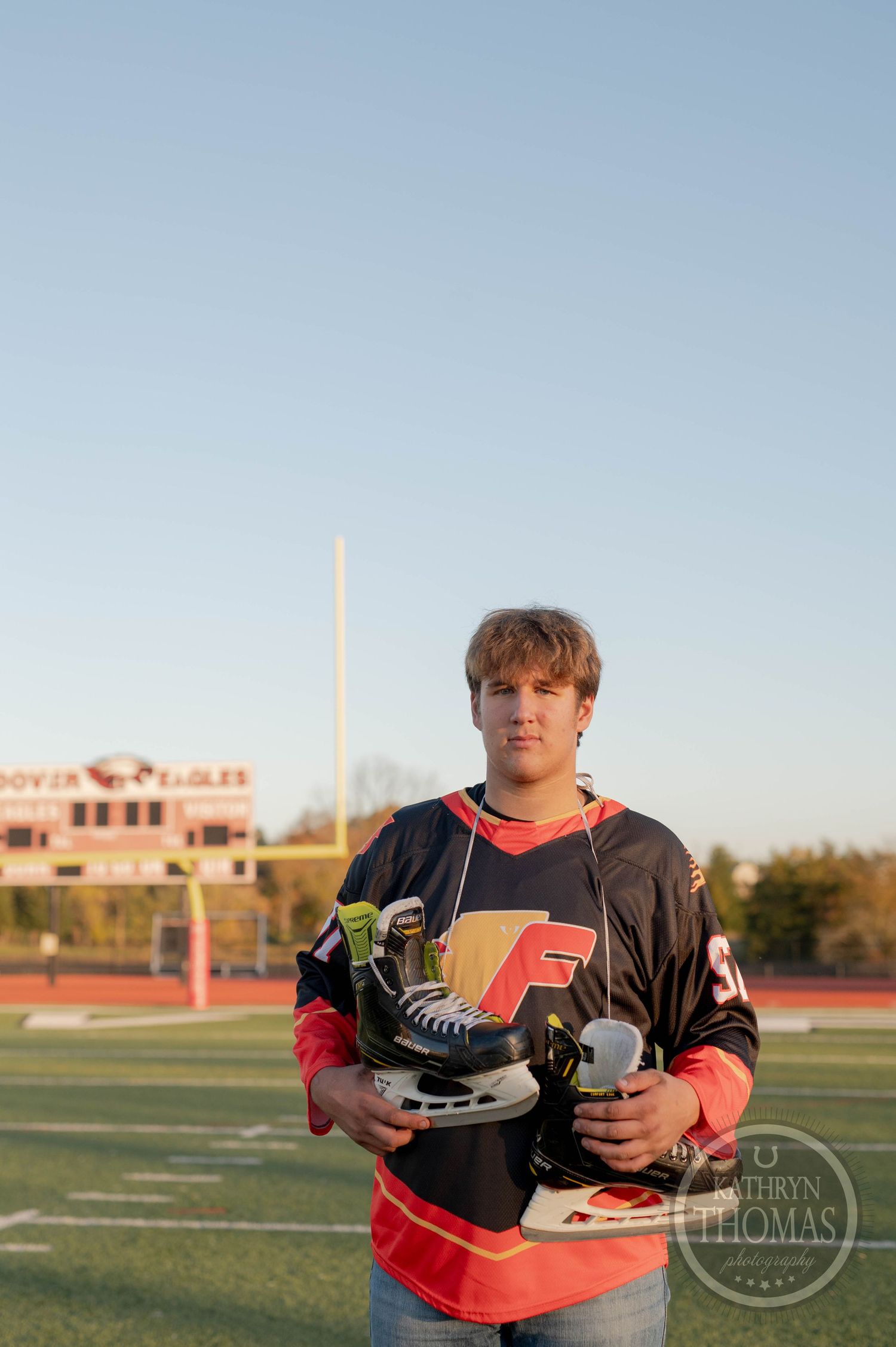 Student athlete in black and red jersey holds hockey skates on football field with stadium in background.