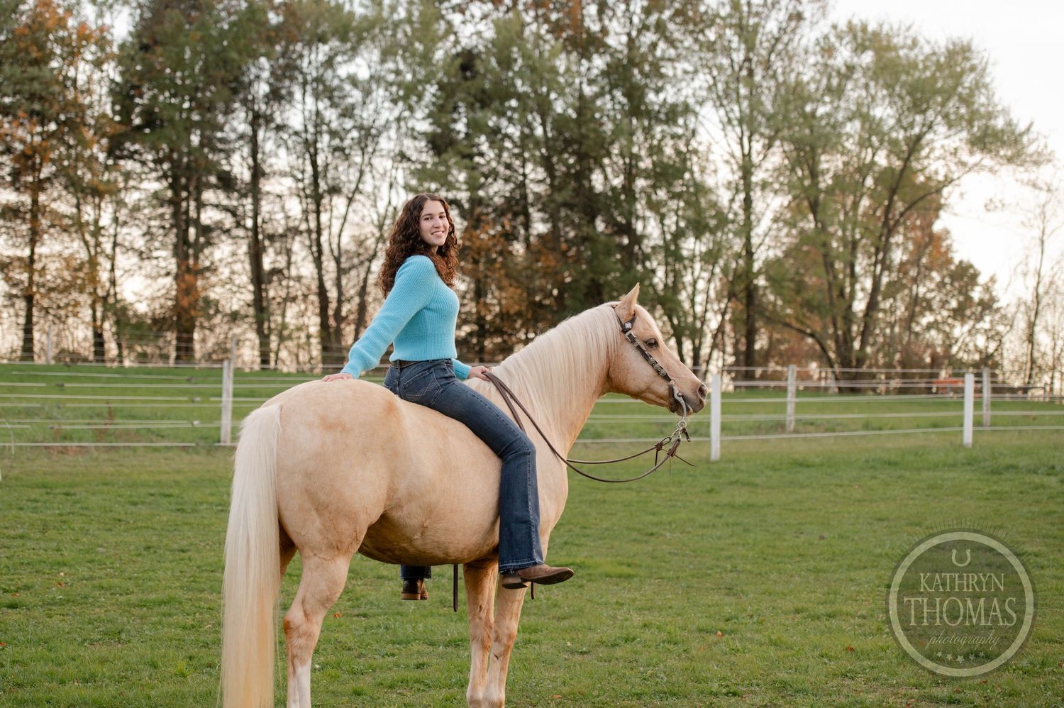 Rider in bright blue sweater horseback riding on palomino horse in fenced pasture.