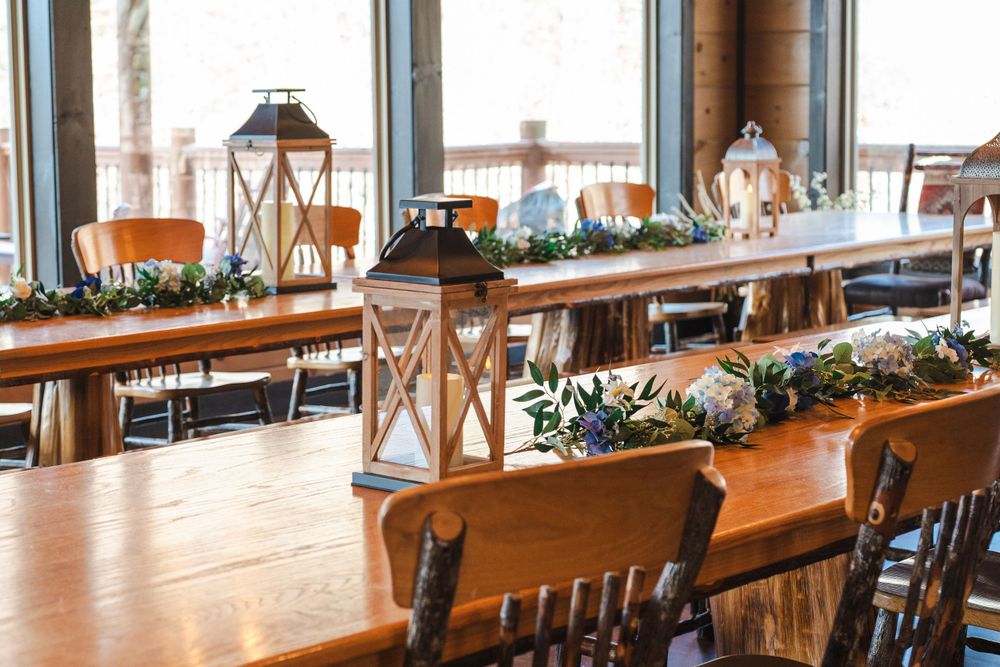 Rustic wooden tables decorated with lanterns and greenery garlands for an elegant wedding reception by large windows.