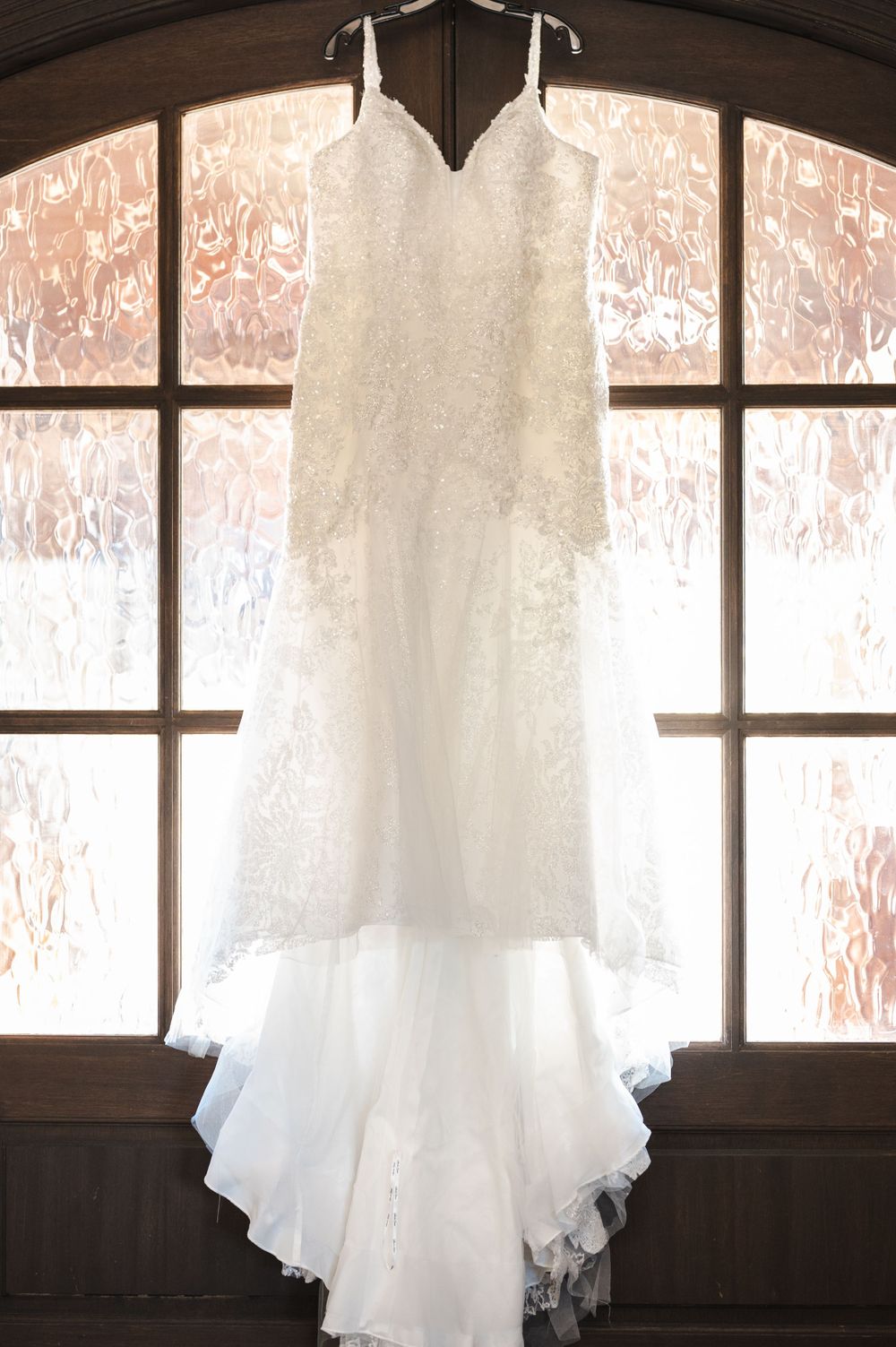 A white lace wedding dress hanging in front of a large arched window with soft natural backlighting.