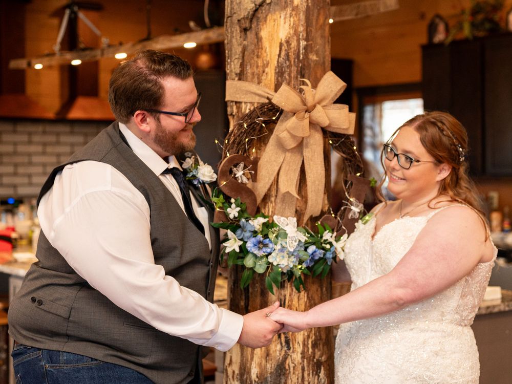 Couple exchanges vows during rustic indoor wedding ceremony with burlap bow and floral decorations on wooden post.