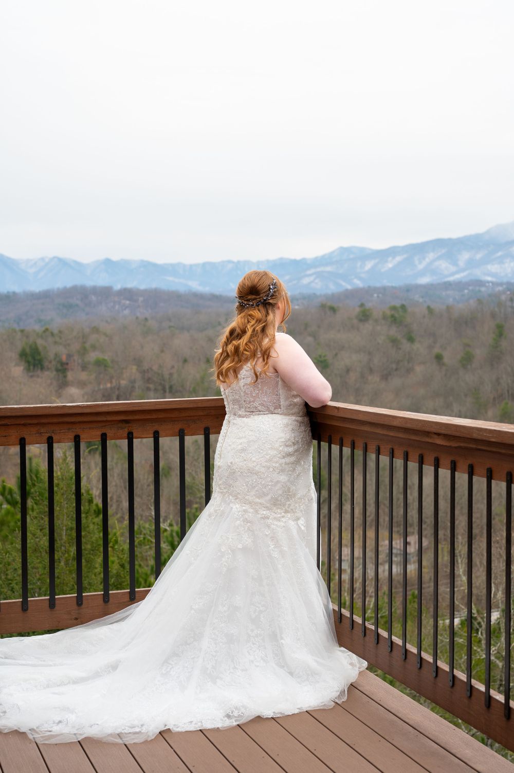 A bride in a lace wedding dress gazes at mountain views from a wooden deck overlooking a scenic valley landscape.