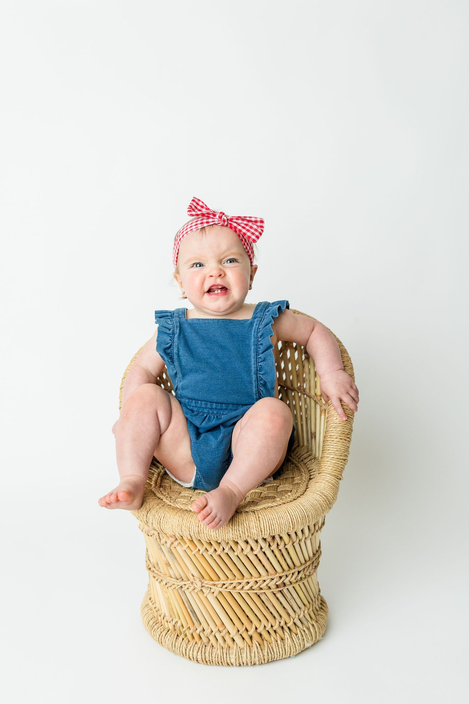 A baby wearing a blue romper and red bow sits in a woven rattan peacock chair against a white background.