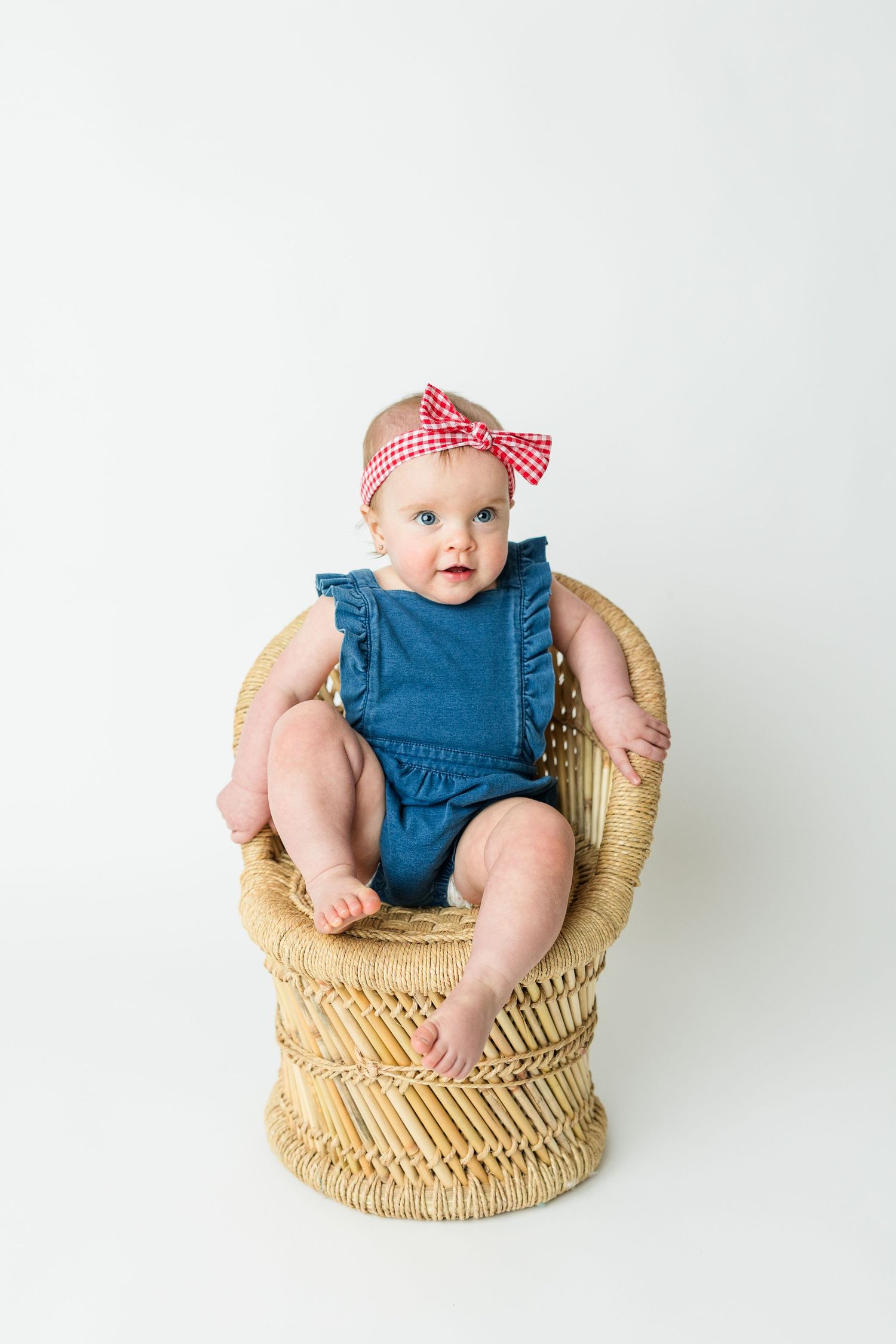 Baby in blue romper with red bow headband sits in woven wicker chair against white background.