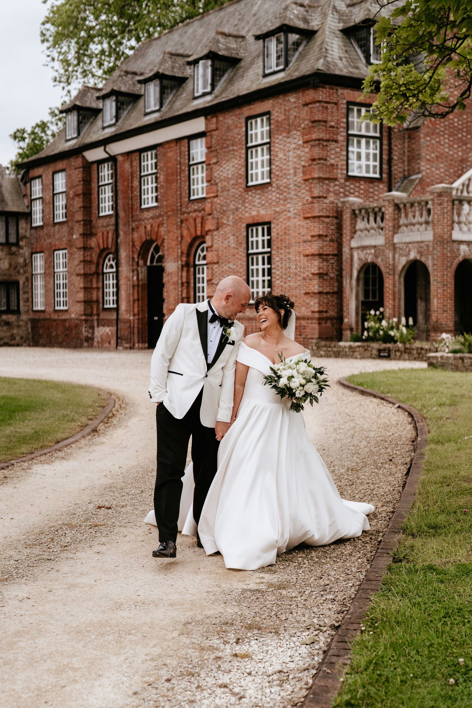 A bride and groom share a romantic moment while walking along a path in front of a grand brick manor house AT SANT FFRAED HOUSE