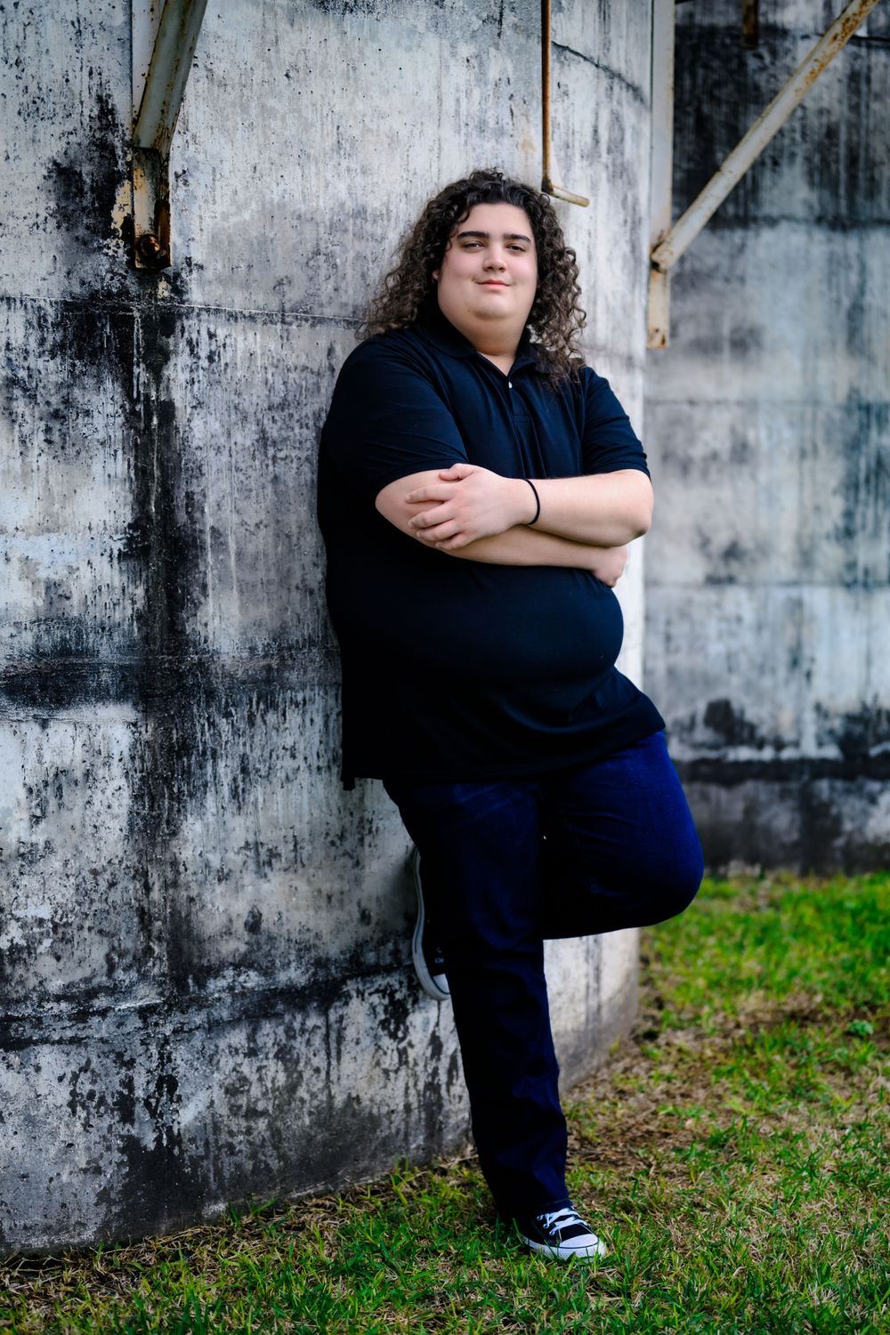 Person with curly dark hair wearing navy blue clothing poses confidently against textured concrete wall outdoors.