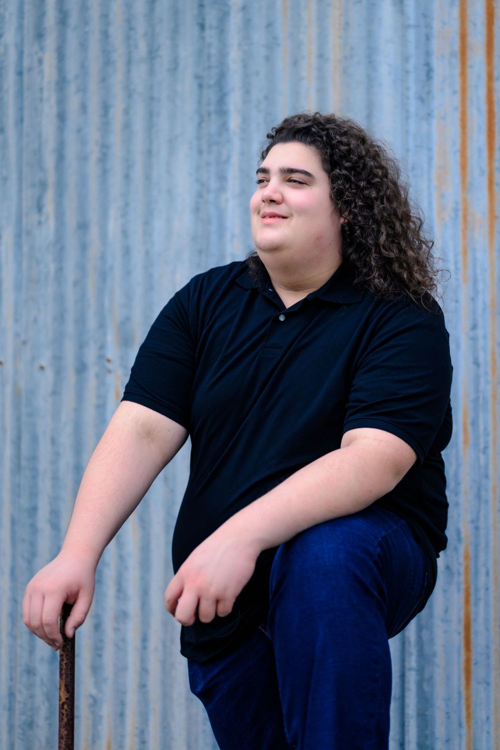 Person with long curly dark hair wearing a black polo shirt and blue pants sits casually against a corrugated metal wall.