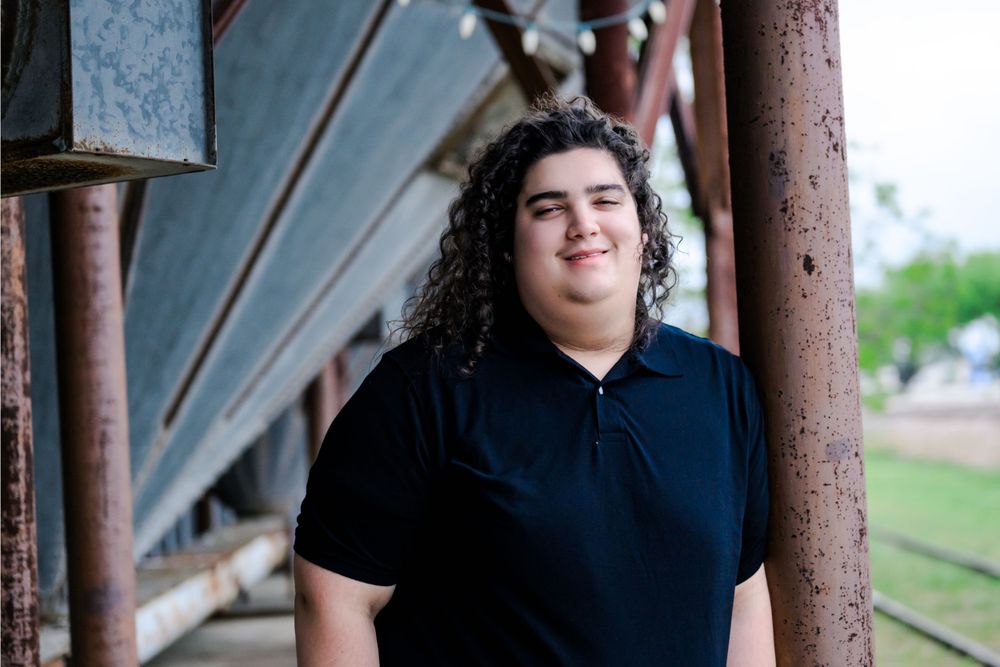 A larger person with curly dark hair wearing a navy polo shirt stands beneath a rustic architectural overhang.