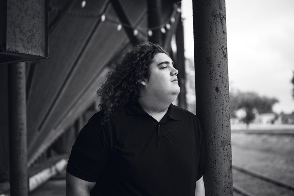 A young person with curly hair stands thoughtfully by a pillar in black and white photography.