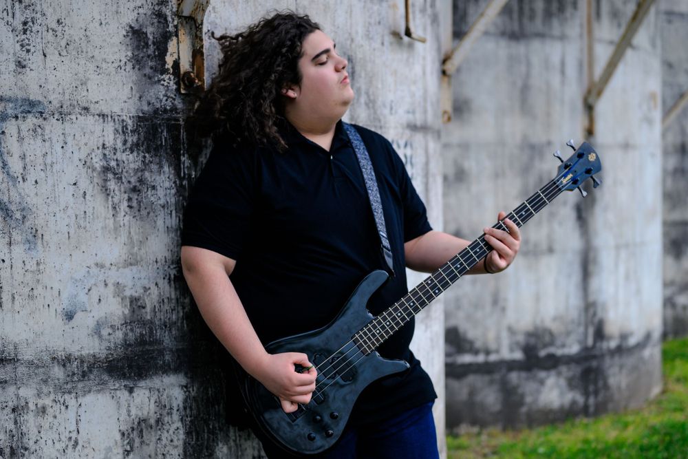 Musician in black clothing playing electric bass guitar while leaning against concrete wall outdoors.