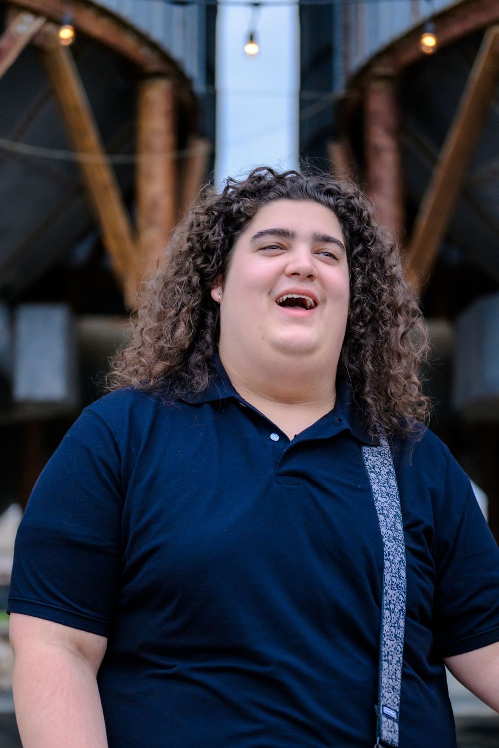 Person with long curly dark hair wearing a navy polo shirt laughs joyfully outdoors.