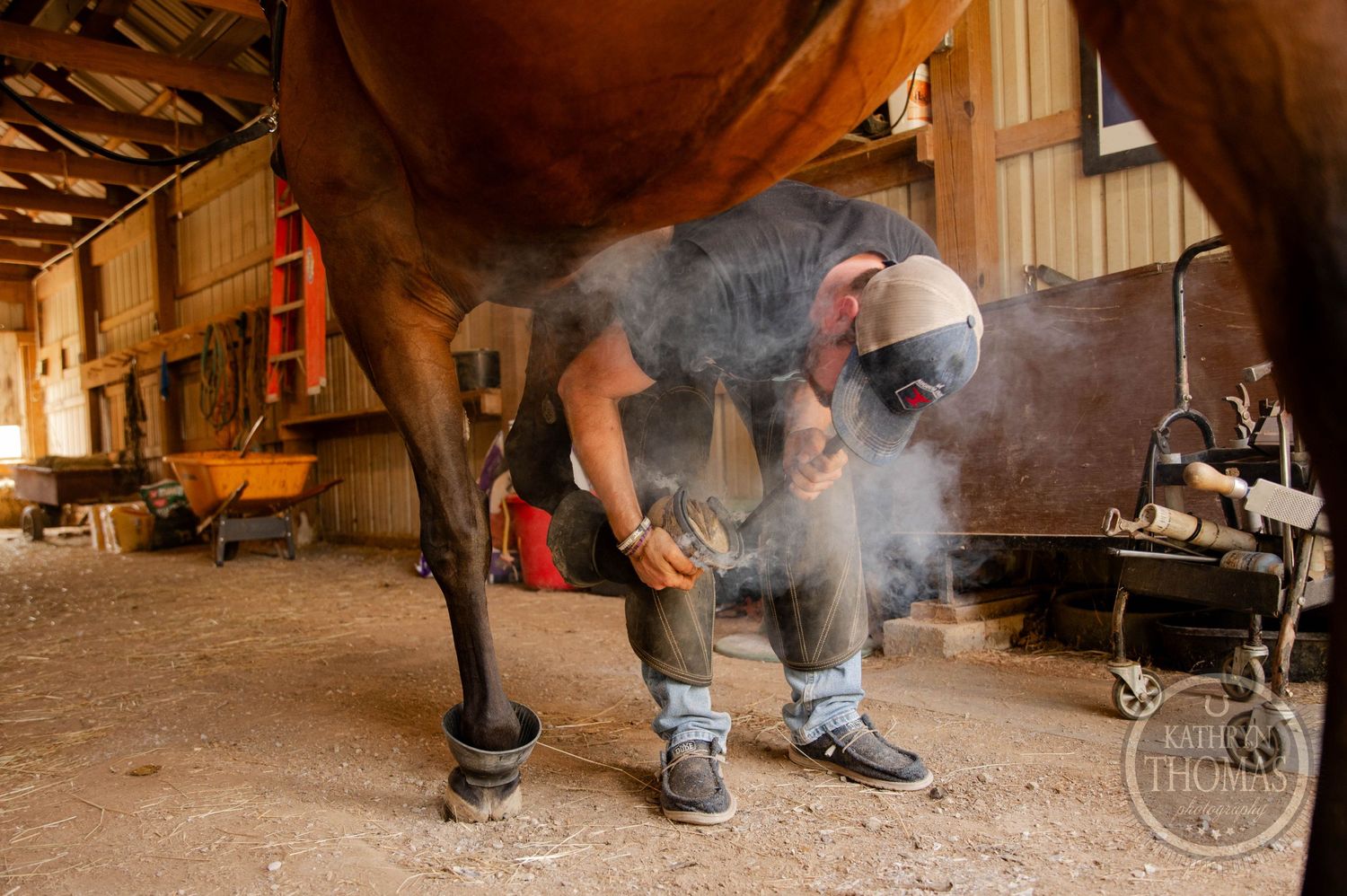 A farrier works underneath a horse in a dimly lit stable with smoke rising from hot metal.