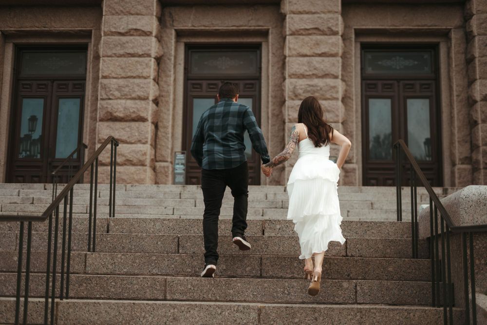 A couple in wedding attire walking up stone steps of a historic building holding hands.