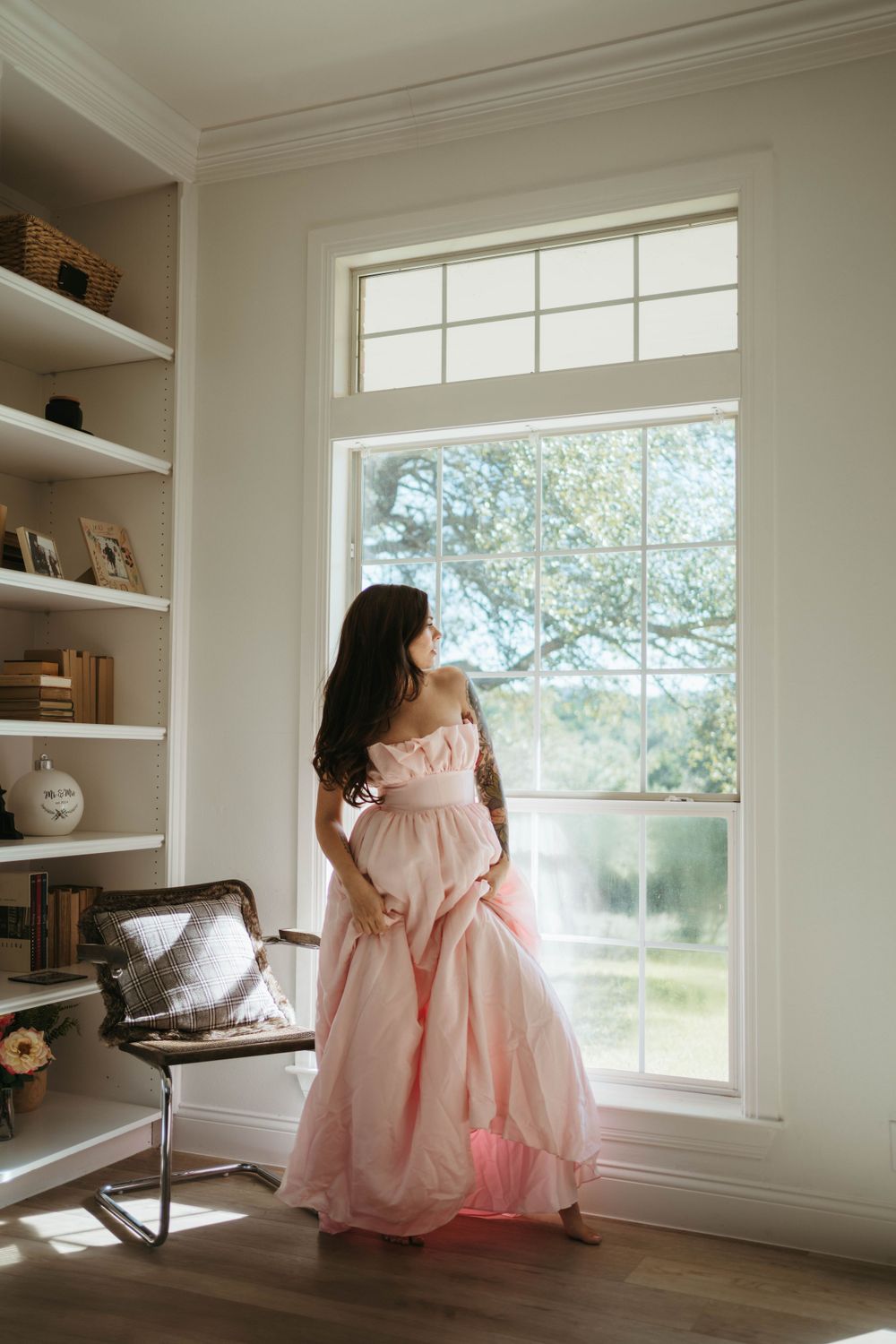 A person in a flowing pink vintage-style dress stands by a large window with built-in shelving in a sunlit room.