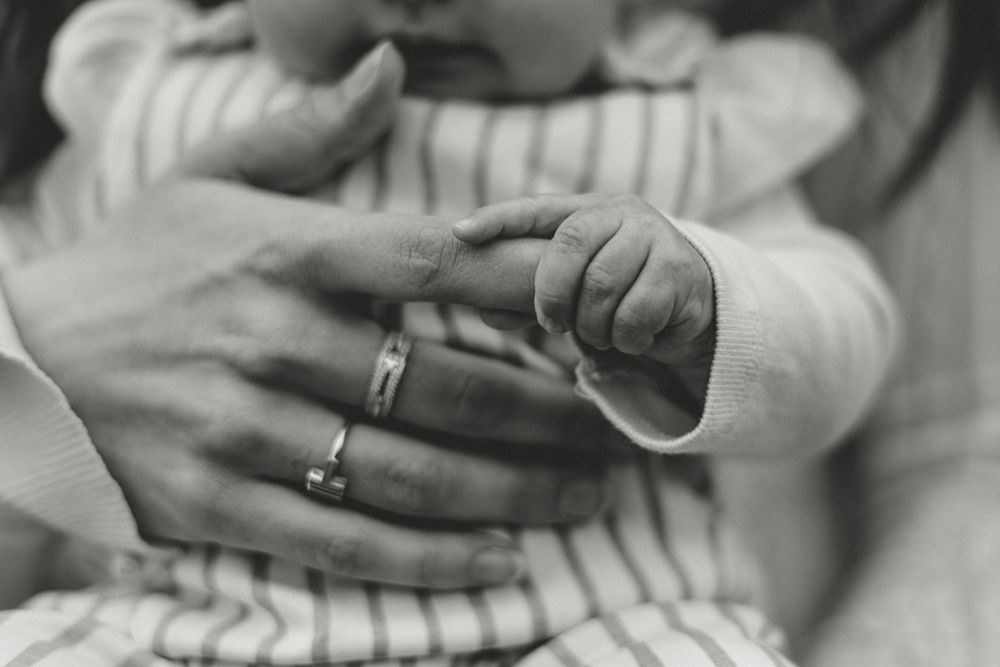 Black and white close-up photograph showing tiny baby fingers grasping an adult hand with wedding rings.
