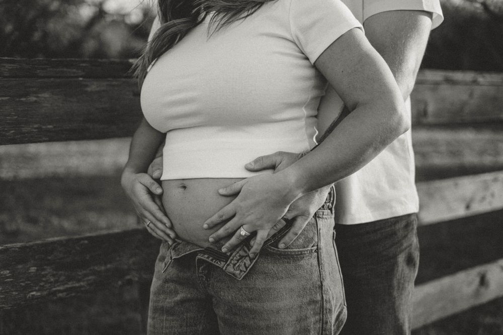 Black and white intimate photo of pregnant belly with hands making heart shape against wooden fence background.