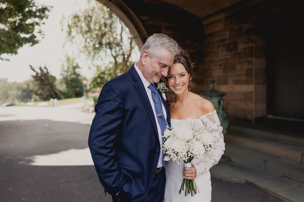 A romantic wedding portrait of a couple embracing outdoors in formal attire with a white floral bouquet.
