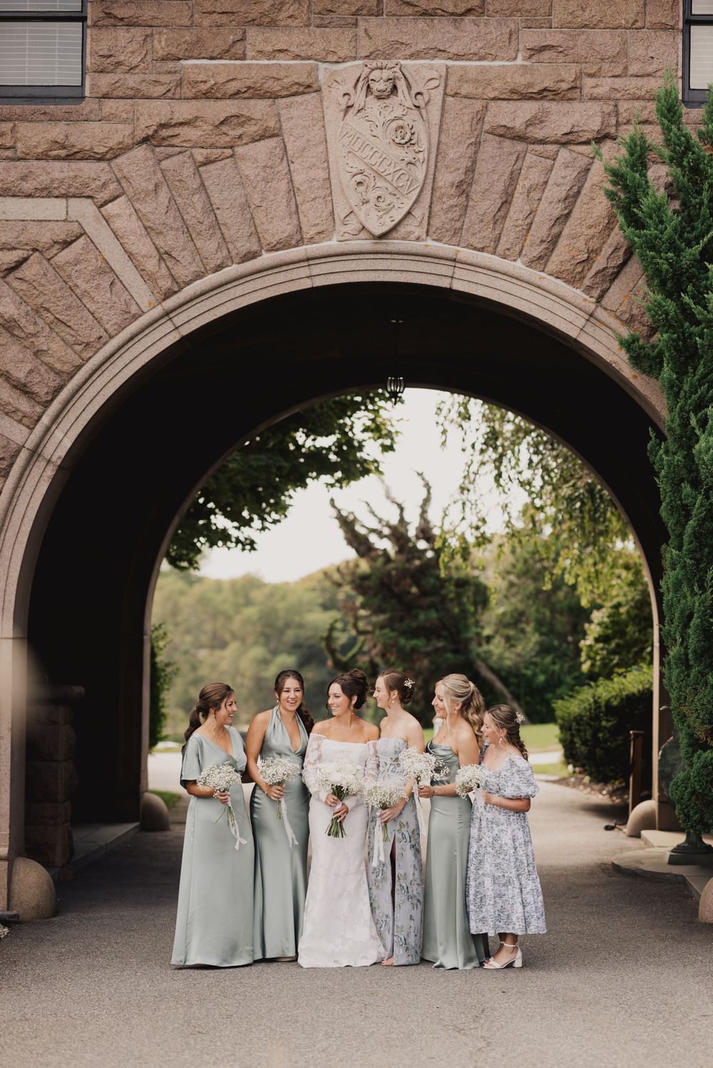 Wedding party poses under a stone archway wearing elegant sage green dresses and holding bouquets.