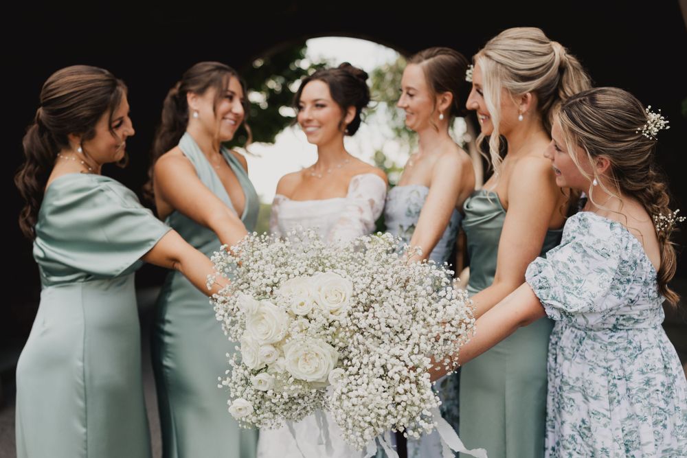 Bridesmaids in sage green dresses gather around a bride holding baby's breath bouquets.