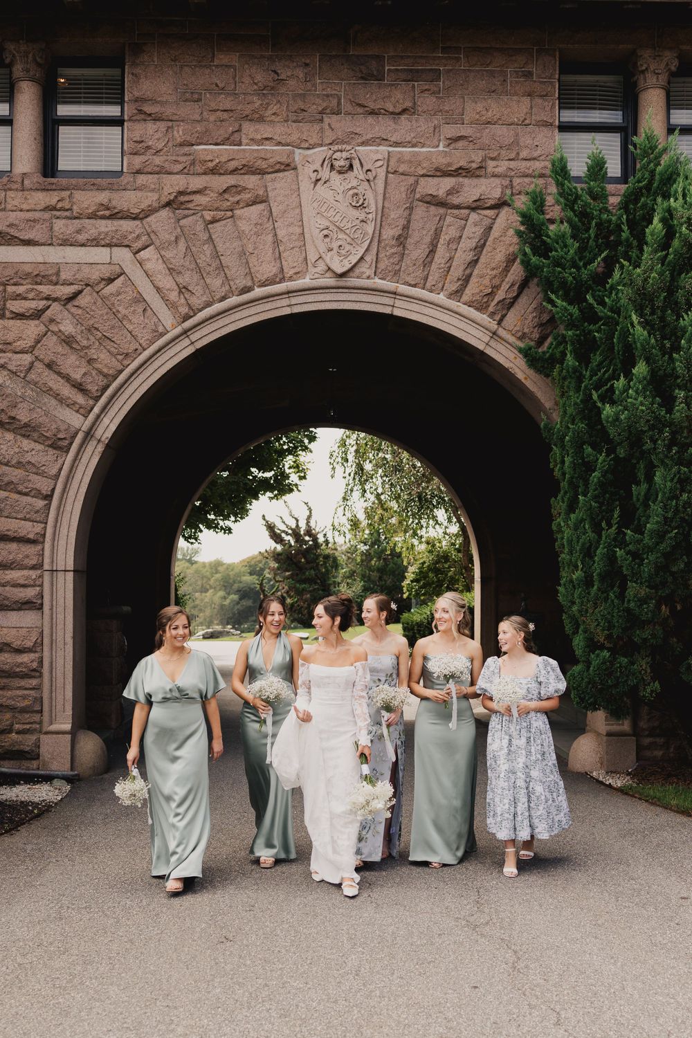 Wedding party walks through stone archway entrance wearing coordinated pastel gowns.