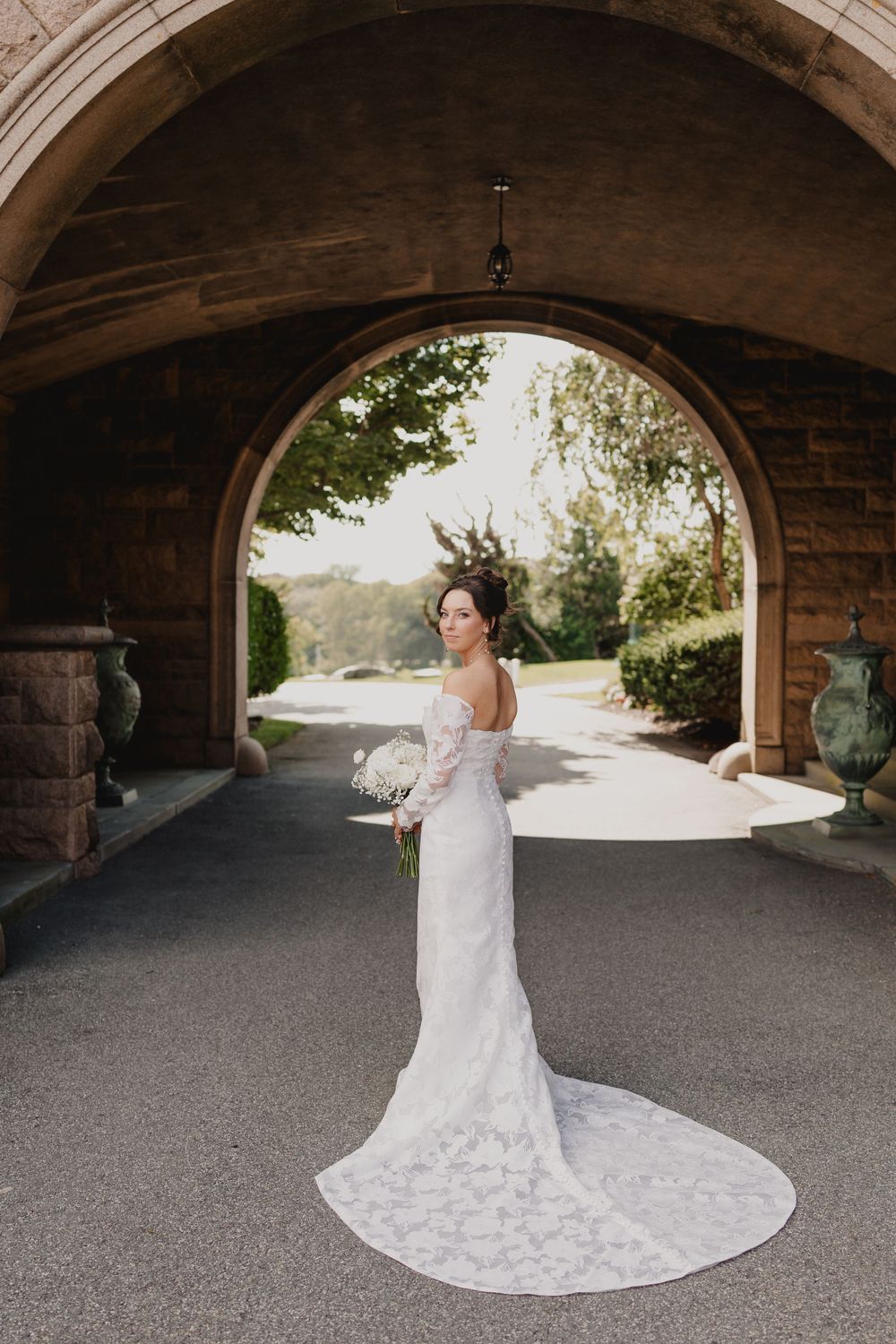 Bride in lace wedding gown with train poses under stone archway.