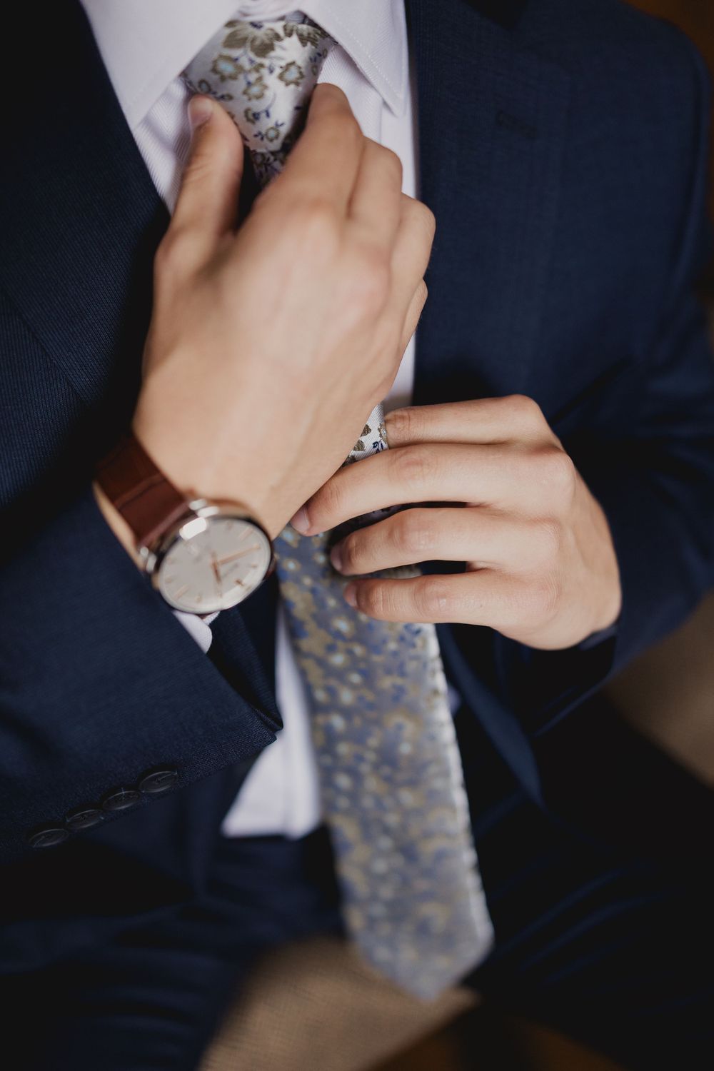 Close-up of hands adjusting gray patterned tie with leather watch visible.