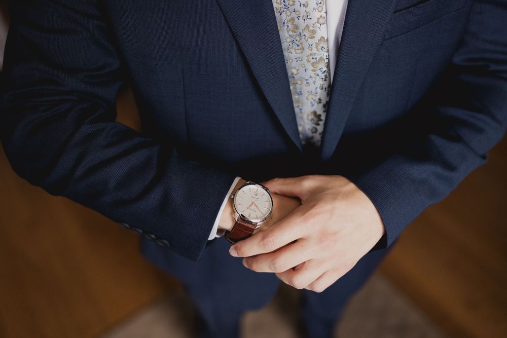 Close-up detail of navy suit with leather watch and hands clasped.