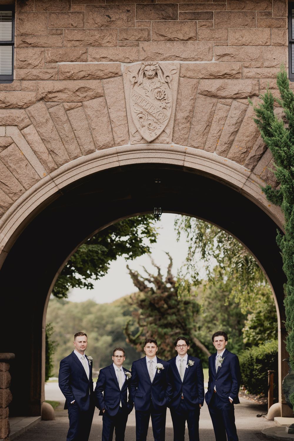 Wedding party poses beneath an ornate stone archway decorated with architectural details.