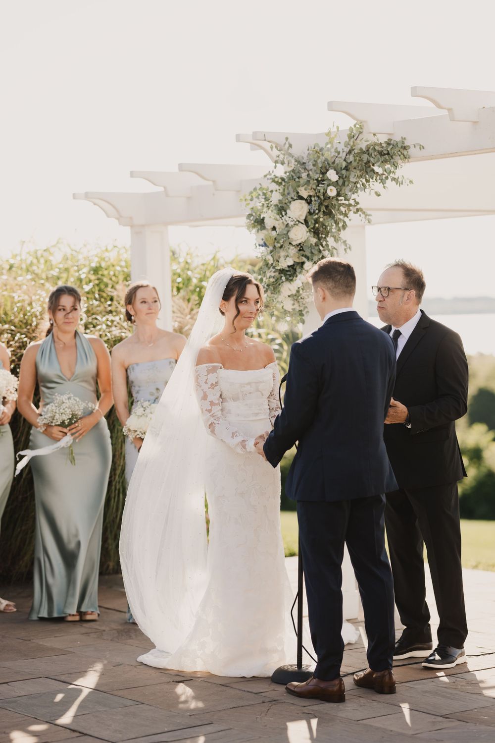 Wedding ceremony under white pergola with floral arrangements and waterfront view.