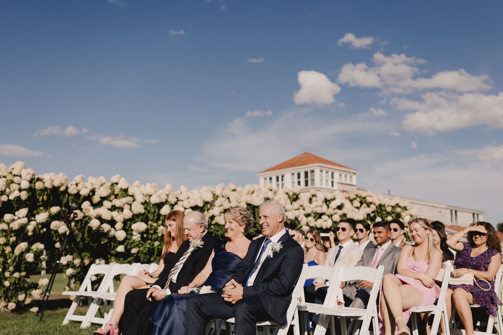 Wedding guests seated in white chairs along floral-lined aisle with historic building in background.