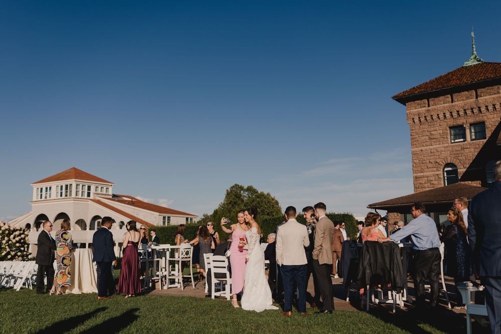 Wedding guests gather on a lawn near a Spanish-style building with tower during an outdoor ceremony at sunset.