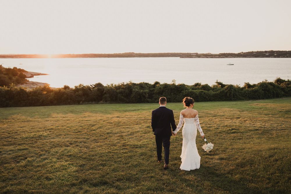 Couple walks on grassy hilltop overlooking waterfront at golden hour.